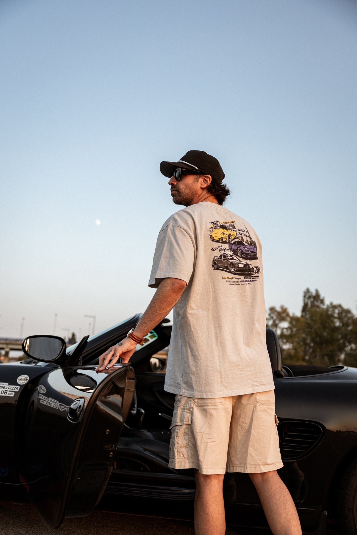 Camiseta Racers Club Midnight Series con estampado trasero de coches, usada por modelo junto a coche descapotable negro