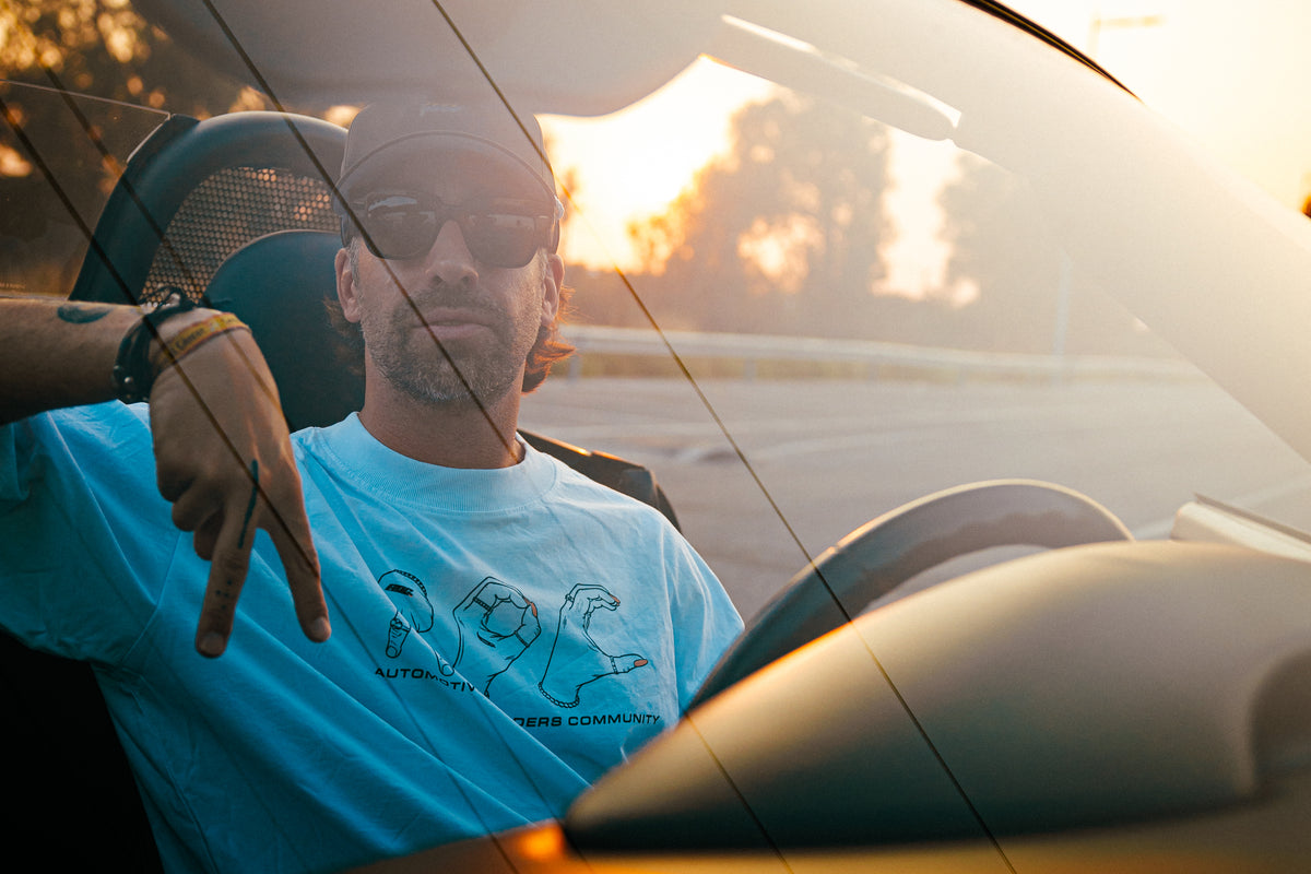Koke Muñoz con camiseta AOC Hands blanca sentado en un Porsche Boxster