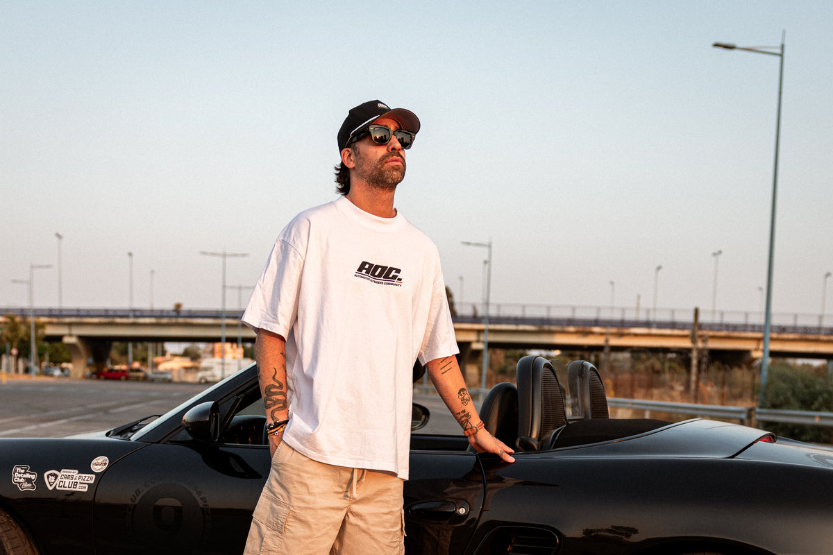 Koke Muñoz con camiseta AOC Logo blanca junto a un Porsche Boxster negro